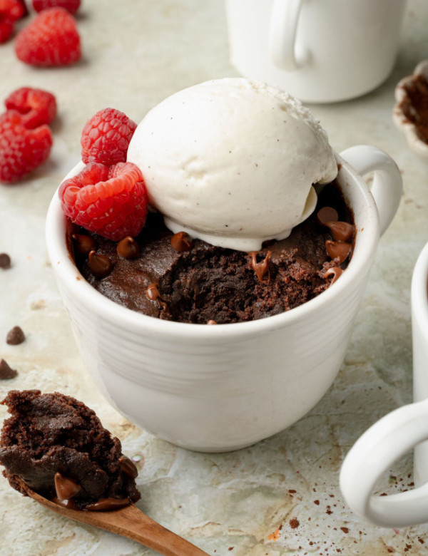 Chocolate cake in a white mug topped with vanilla ice cream and fresh raspberries. A spoonful of moist chocolate cake lies beside the mug on a marble surface.