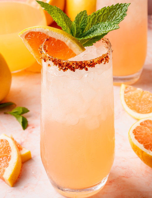 Close-up of a single glass of Paloma cocktail topped with crushed ice, garnished with a grapefruit slice, lemon wedge, and mint sprig. The glass is rimmed with chili-lime salt and placed on a light pink marble background.