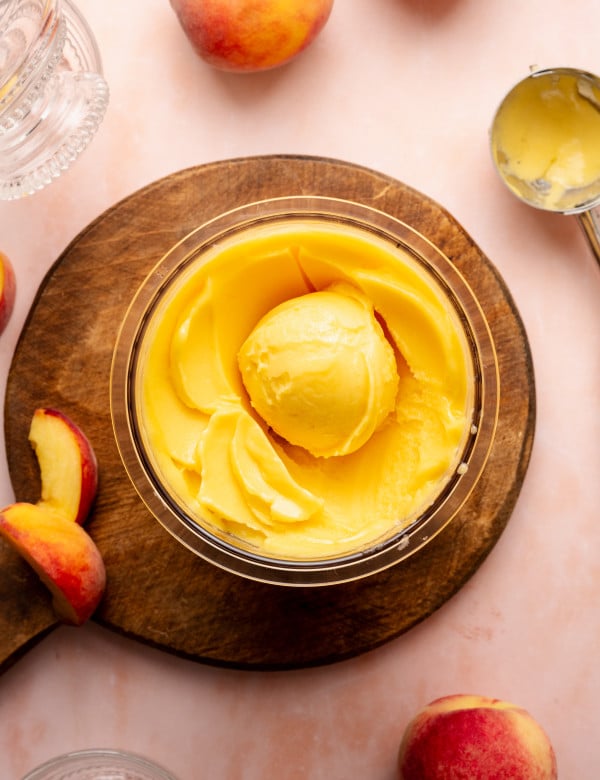 Peach sorbet in ninja creami pint container scooped in container on top of wood cutting board with peaches on the side of the board and on the table.