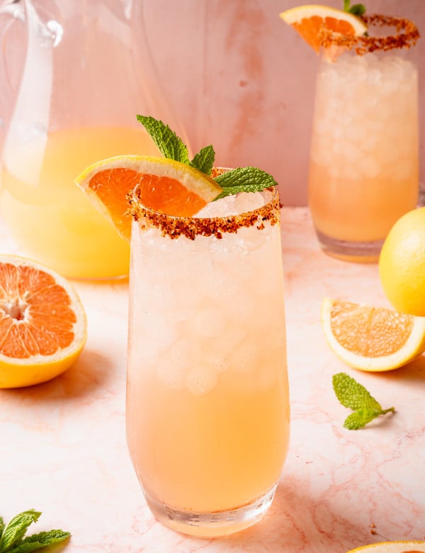 Close-up of a single glass of Paloma cocktail topped with crushed ice, garnished with a grapefruit slice, lemon wedge, and mint sprig. The glass is rimmed with chili-lime salt and placed on a light pink marble background.