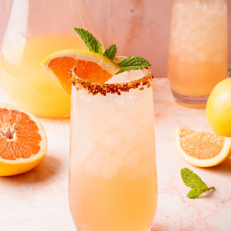 Close-up of a single glass of Paloma cocktail topped with crushed ice, garnished with a grapefruit slice, lemon wedge, and mint sprig. The glass is rimmed with chili-lime salt and placed on a light pink marble background.
