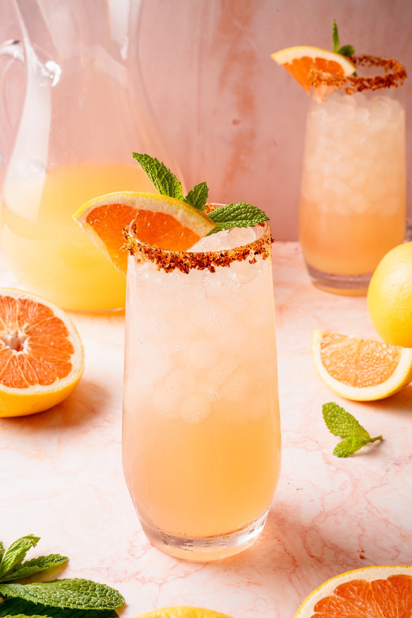 Close-up of a single glass of Paloma cocktail topped with crushed ice, garnished with a grapefruit slice, lemon wedge, and mint sprig. The glass is rimmed with chili-lime salt and placed on a light pink marble background.