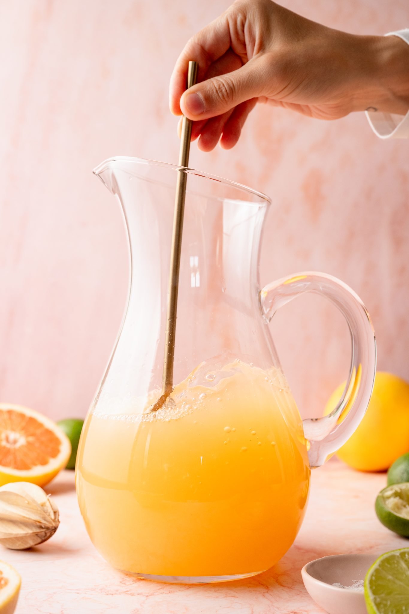A hand stirs a large clear pitcher of freshly mixed Paloma cocktail with a golden stirring stick. Cut citrus and cocktail ingredients are scattered around the pitcher on a pink background.