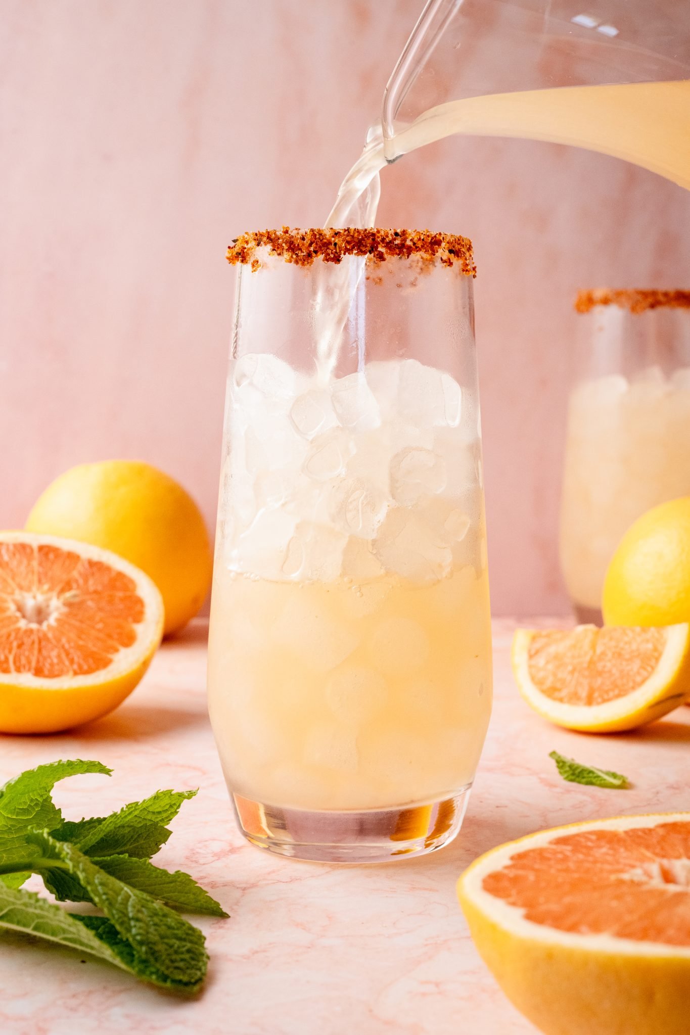 A glass with a chili-lime rim being filled with grapefruit Paloma cocktail over ice from a clear pitcher. The background includes sliced grapefruit, lemons, and mint leaves on a pink marble surface.