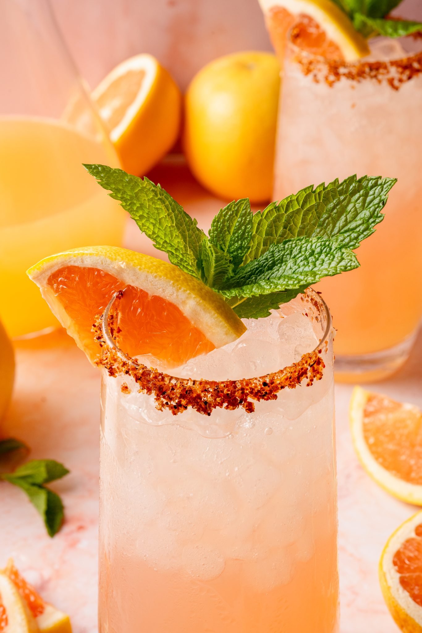 Close-up of a single glass of Paloma cocktail topped with crushed ice, garnished with a grapefruit slice, lemon wedge, and mint sprig. The glass is rimmed with chili-lime salt and placed on a light pink marble background.