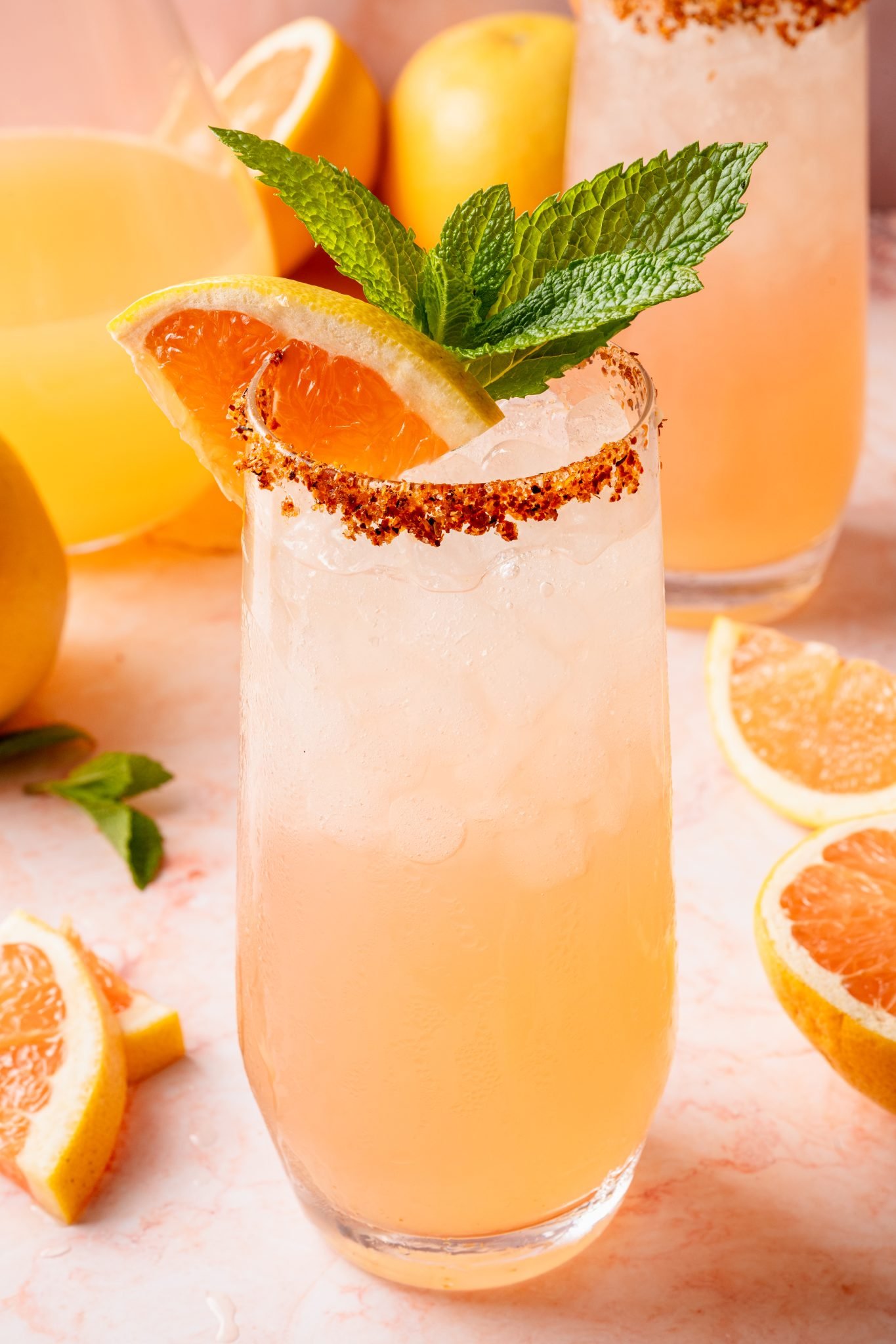 A tall glass of pink Paloma cocktail filled with crushed ice, garnished with a grapefruit wedge, lemon slice, and fresh mint, with a chili-lime salt rim. A pitcher of Paloma and another garnished glass are visible in the background alongside citrus fruits and mint on a pink marble surface.