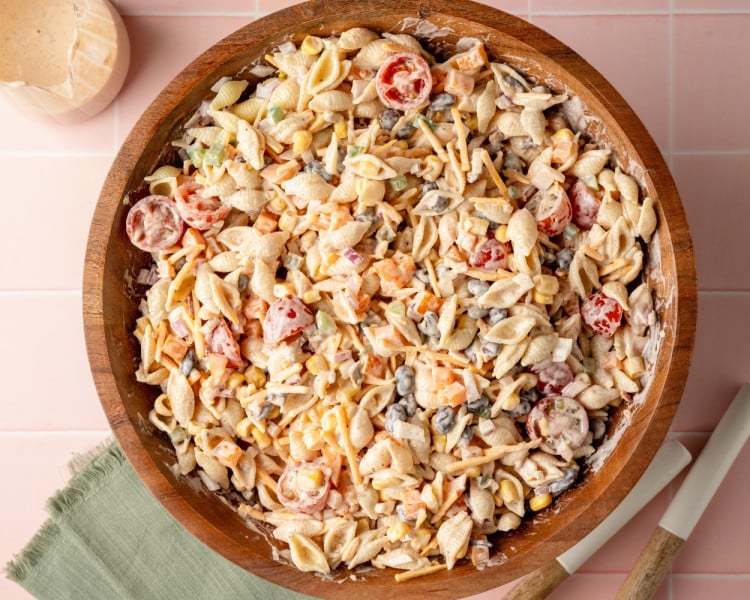 Overhead view of fully mixed creamy taco pasta salad in a large wooden bowl. Shell pasta, cherry tomatoes, corn, black beans, shredded cheese, and diced veggies are coated in the creamy taco dressing.
