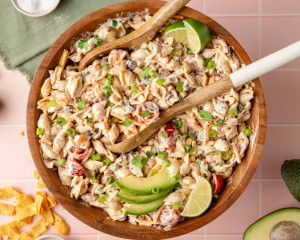 Finished taco pasta salad served in a wooden bowl, garnished with avocado slices, lime wedges, chopped green onions, and fresh cilantro. Wooden utensils rest in the bowl and ingredients like chips and salt are scattered around the pink tiled background.