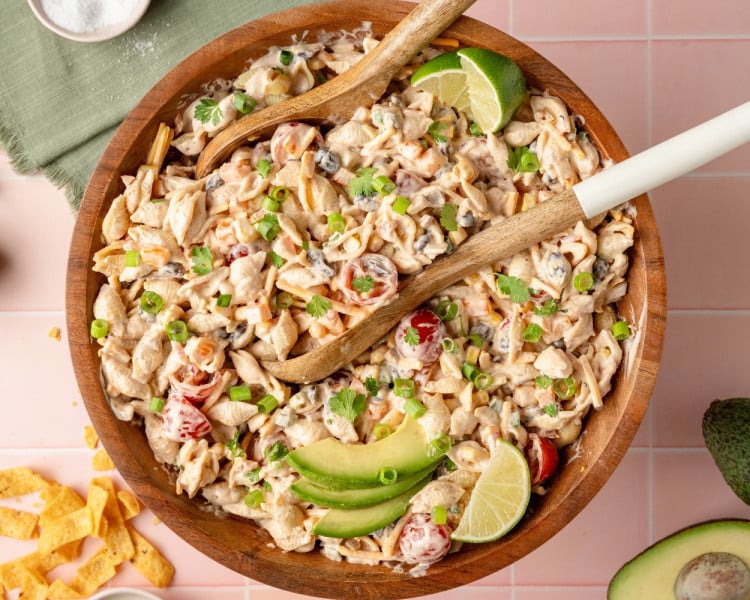 Finished taco pasta salad served in a wooden bowl, garnished with avocado slices, lime wedges, chopped green onions, and fresh cilantro. Wooden utensils rest in the bowl and ingredients like chips and salt are scattered around the pink tiled background.