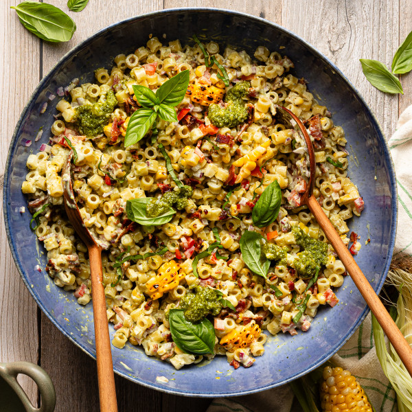 Pesto pasta salad tossed together in large blue serving bowl with wood spoons and garnished with fresh basil.