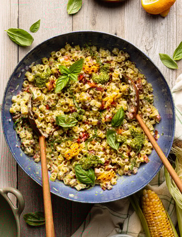 Pesto pasta salad tossed together in large blue serving bowl with wood spoons and garnished with fresh basil.
