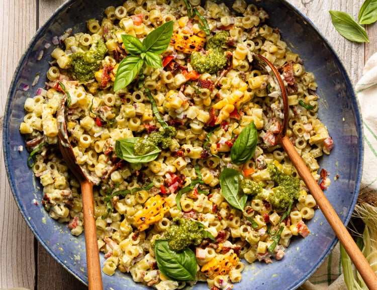 Pesto pasta salad tossed together in large blue serving bowl with wood spoons and garnished with fresh basil.