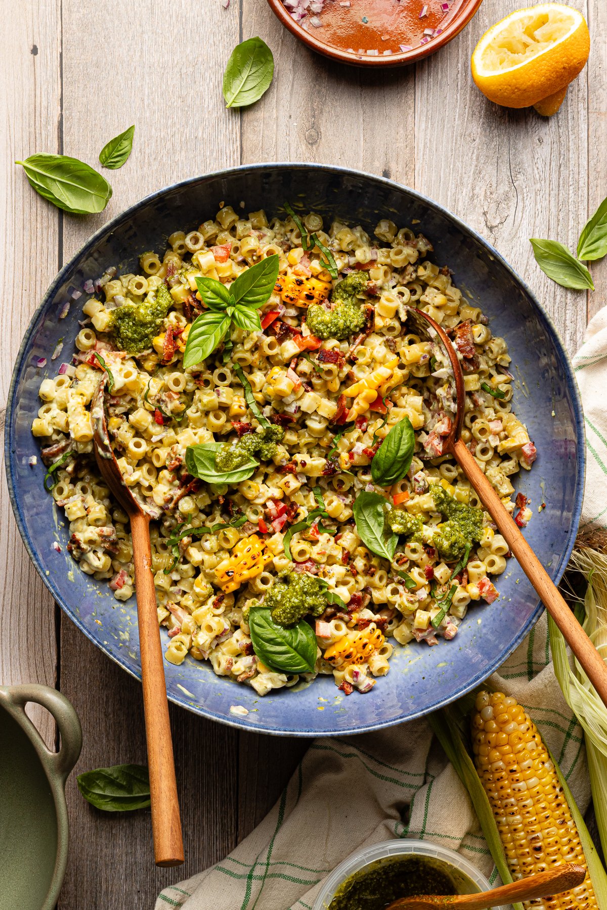 Pesto pasta salad tossed together in large blue serving bowl with wood spoons and garnished with fresh basil.