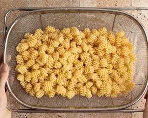 Cooked mafaldine pasta draining in a large metal colander over a tiled countertop. Hands are holding the colander from either side.