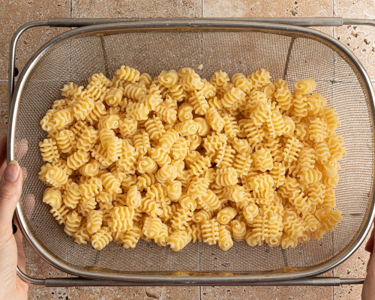 Cooked mafaldine pasta draining in a large metal colander over a tiled countertop. Hands are holding the colander from either side.