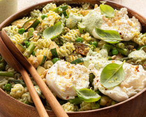 Close-up side view of finished green goddess pasta salad in a wooden bowl with burrata, fresh basil, pine nuts, and a drizzle of green dressing. Wooden salad servers rest inside the bowl.