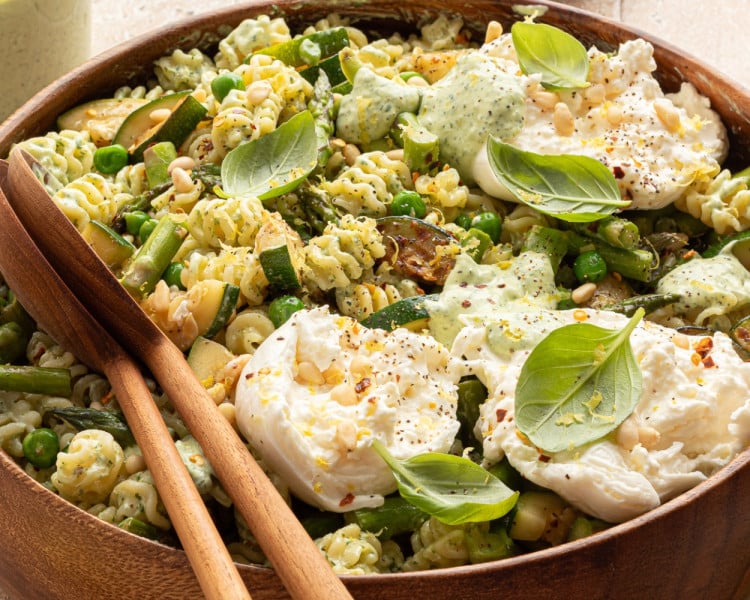 Close-up side view of finished green goddess pasta salad in a wooden bowl with burrata, fresh basil, pine nuts, and a drizzle of green dressing. Wooden salad servers rest inside the bowl.