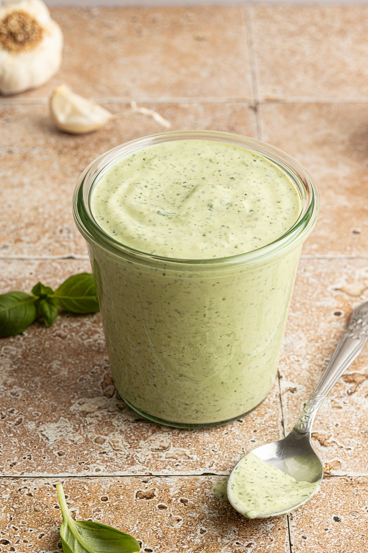 A glass jar filled with creamy green goddess dressing sits on a tile surface, with a spoonful of the dressing resting nearby. Fresh basil leaves and a head of garlic are blurred in the background.