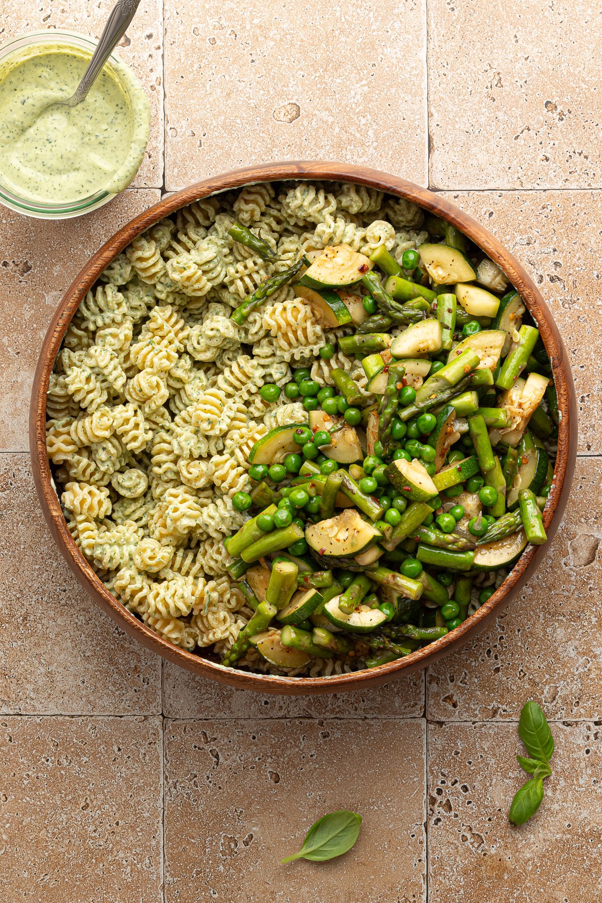 Overhead view of a large wooden bowl filled halfway with green goddess pasta and halfway with sautéed vegetables including zucchini, asparagus, and peas. A jar of creamy green herb dressing sits nearby on a tiled surface.