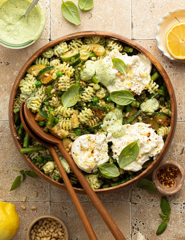 Overhead view of a finished green goddess pasta salad topped with dollops of burrata cheese, pine nuts, and fresh basil. The salad is served in a large wooden bowl surrounded by ingredients like lemon, red pepper flakes, and green herb dressing.