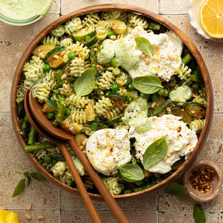 Overhead view of a finished green goddess pasta salad topped with dollops of burrata cheese, pine nuts, and fresh basil. The salad is served in a large wooden bowl surrounded by ingredients like lemon, red pepper flakes, and green herb dressing.