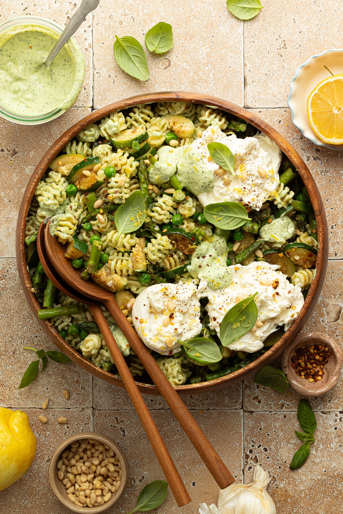 Overhead view of a finished green goddess pasta salad topped with dollops of burrata cheese, pine nuts, and fresh basil. The salad is served in a large wooden bowl surrounded by ingredients like lemon, red pepper flakes, and green herb dressing.