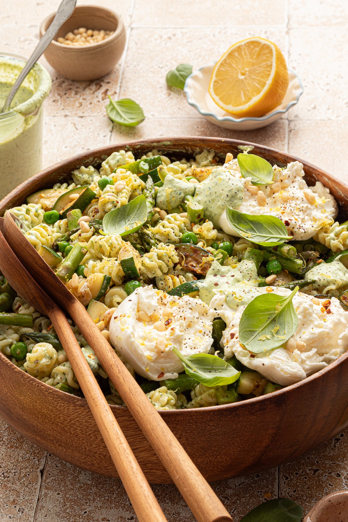 Angled close-up of a green goddess pasta salad in a wooden bowl, garnished with creamy burrata, green dressing, pine nuts, and fresh basil. Wooden salad servers rest in the bowl, with lemon and basil leaves nearby.