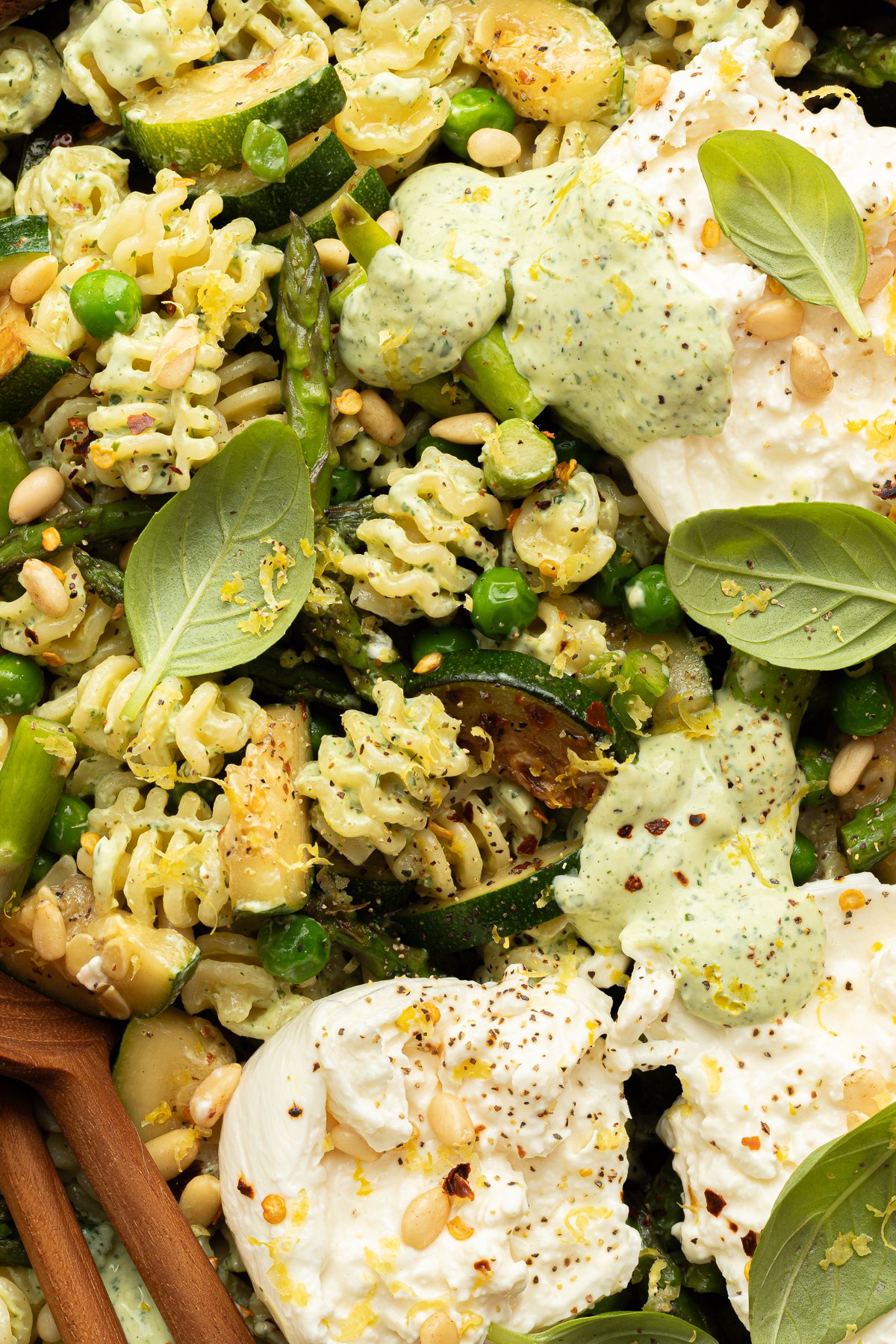Extreme close-up of the pasta salad highlighting spiral pasta coated in green goddess dressing, tender vegetables, pine nuts, and creamy burrata topped with cracked pepper and lemon zest.