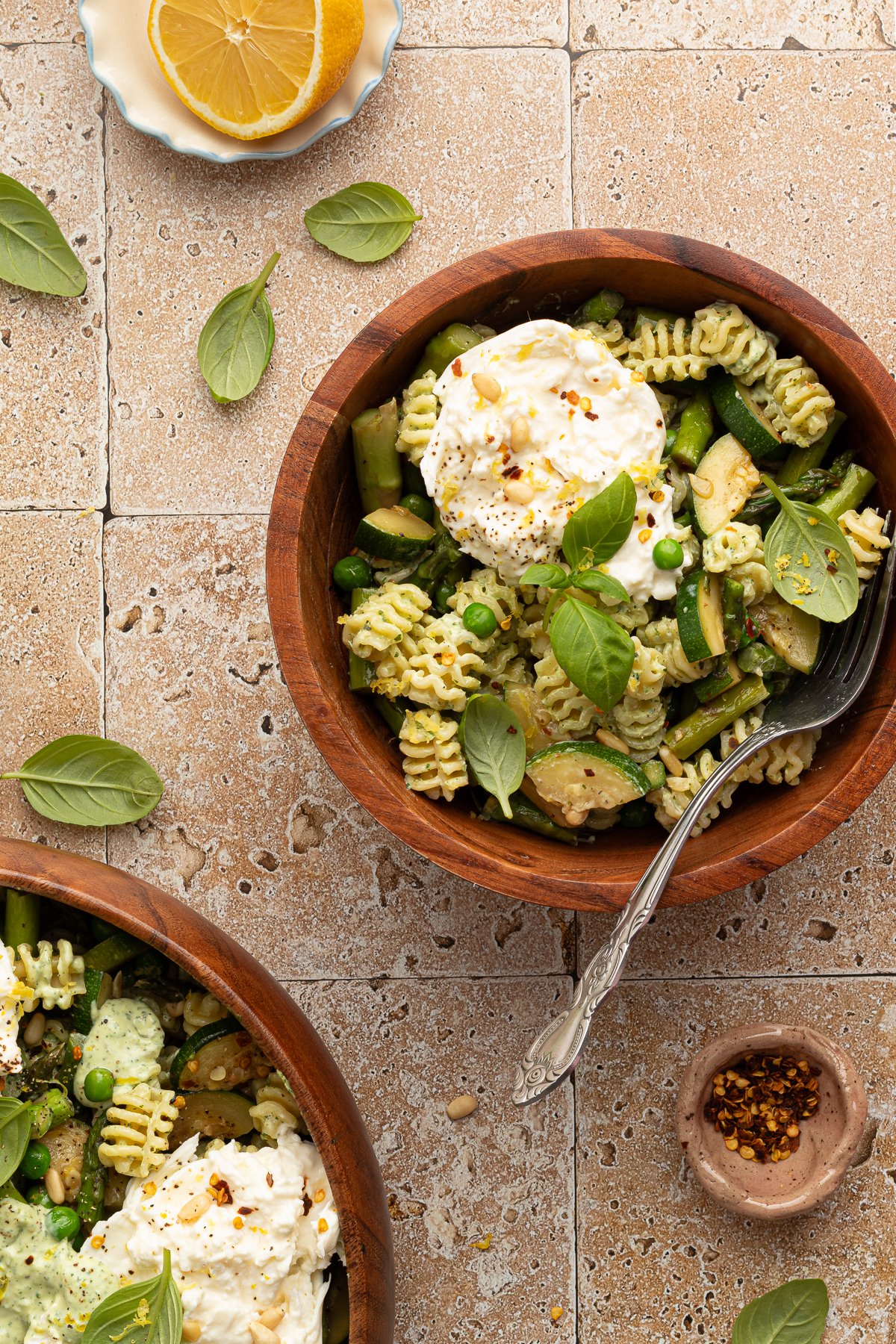 Overhead view of a small wooden bowl filled with green goddess pasta salad and a scoop of burrata cheese, garnished with basil and pine nuts. A larger bowl of pasta is partially visible nearby, with lemon slices and seasonings scattered around.