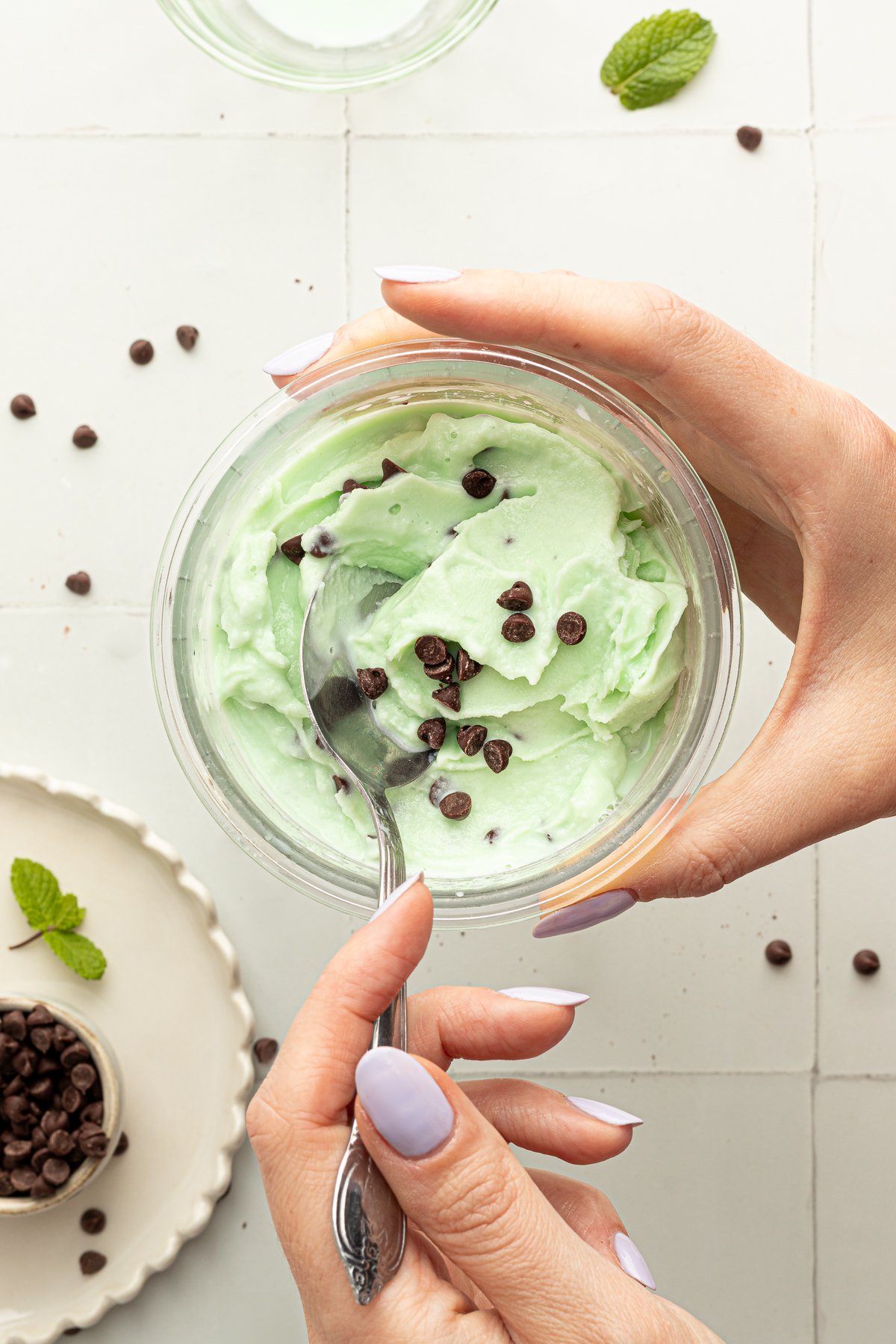 Hands holding a pint of mint chocolate chip Ninja CREAMi protein ice cream, scooping a bite with a spoon, chocolate chips scattered across the surface.