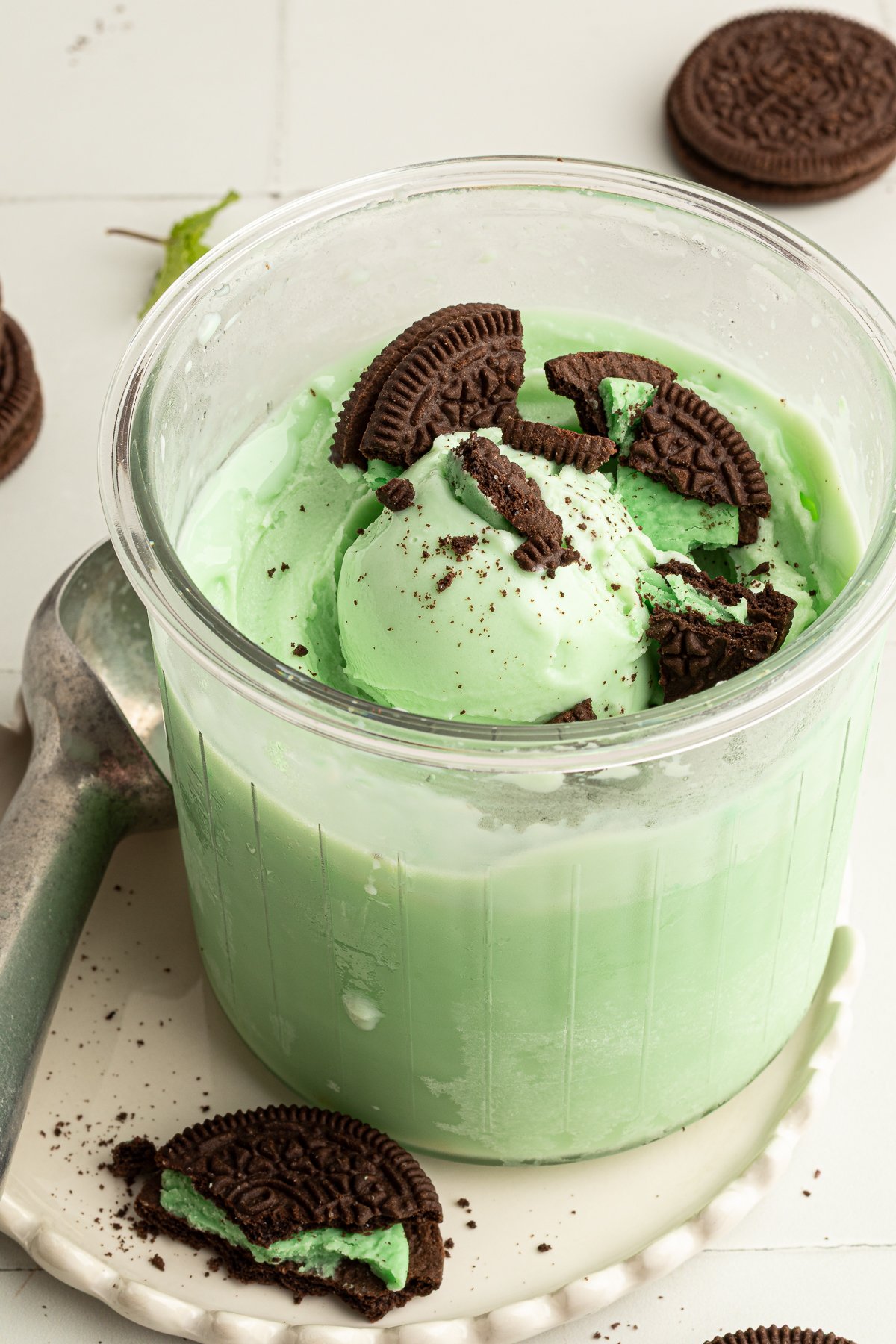 Ninja CREAMi pint of mint chocolate chip protein ice cream topped with broken mint sandwich cookies, served with an ice cream scoop and mint leaves nearby.