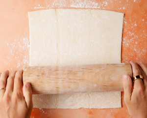 Close-up of puff pastry being rolled out on a floured surface with both hands on the rolling pin. This step is preparing the top lattice crust.
