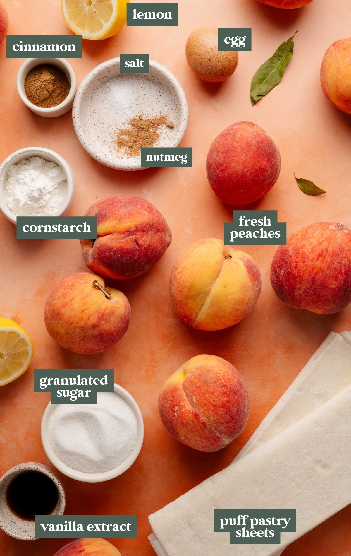 Flat lay of labeled ingredients for peach slab pie on a peach-colored background. Items include fresh whole peaches, puff pastry sheets, a bowl of granulated sugar, a dish of cornstarch, cinnamon, nutmeg and salt in a speckled bowl, a halved lemon, a whole egg, vanilla extract, and a couple of green leaves scattered among the ingredients.