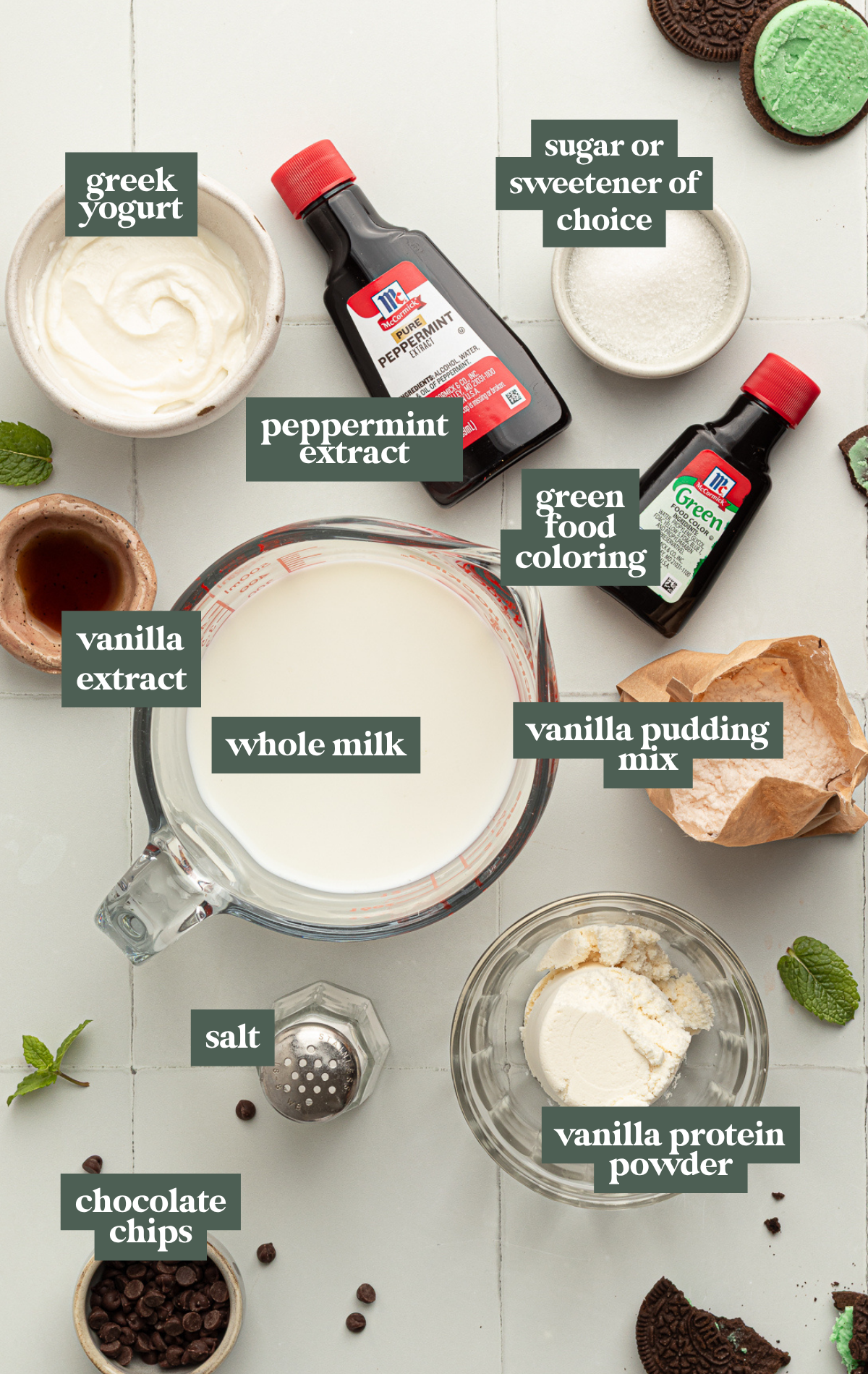 Overhead view of mint chocolate chip Ninja CREAMi ice cream ingredients arranged on a white tile surface, including milk, peppermint extract, green food coloring, pudding mix, protein powder, mini chocolate chips, and mint sandwich cookies.