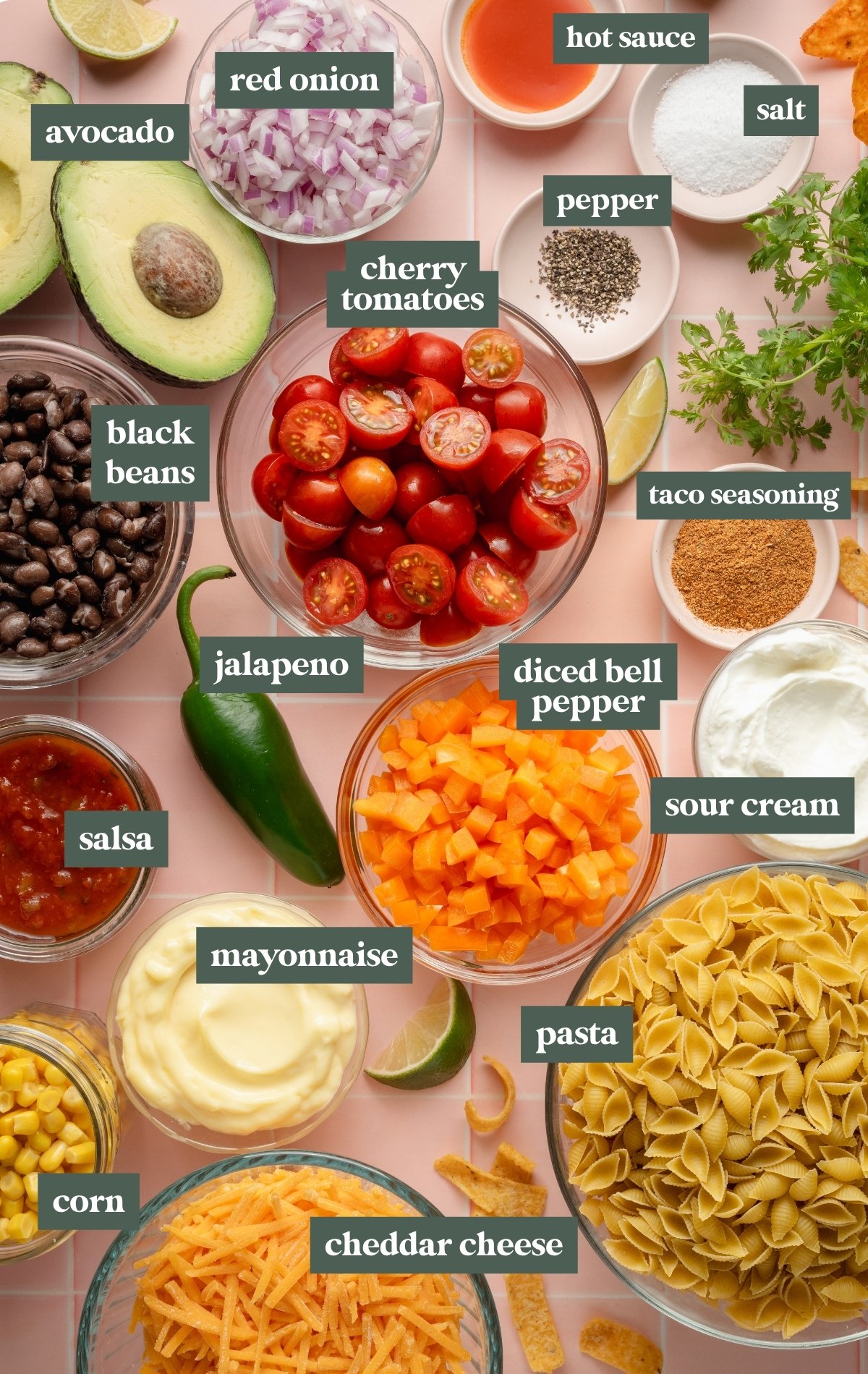 Flat lay of labeled ingredients for taco pasta salad arranged on a pink tile surface. Ingredients include avocado, red onion, cherry tomatoes, black beans, jalapeño, diced bell pepper, salsa, corn, mayonnaise, sour cream, cheddar cheese, pasta shells, taco seasoning, salt, pepper, hot sauce, and fresh cilantro.