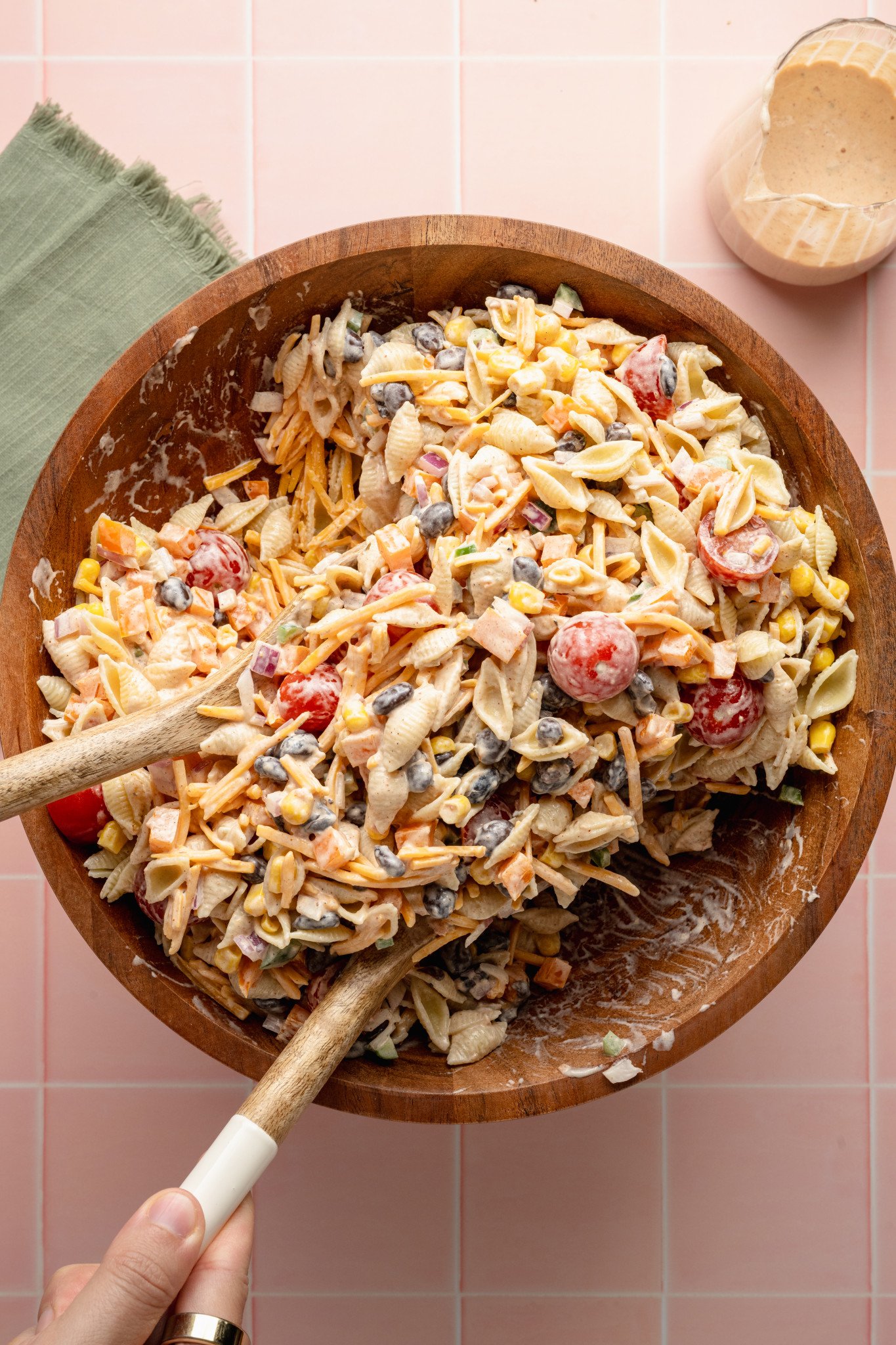 Top-down view of taco pasta salad being mixed with wooden utensils in a large wooden bowl. The creamy salad includes shell pasta, cherry tomatoes, black beans, corn, shredded cheese, diced peppers, and onions, coated in a taco-seasoned dressing.