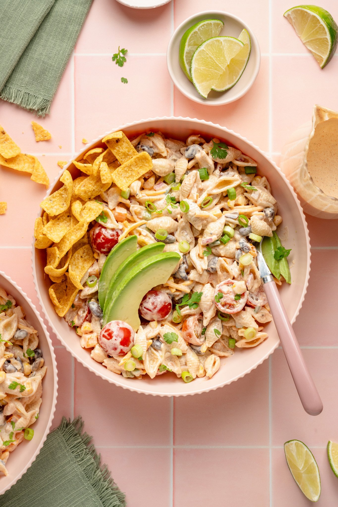 Top-down view of a bowl of taco pasta salad topped with avocado slices, green onions, and cilantro. Fritos are arranged on the side of the bowl, and lime wedges and extra dressing are visible nearby on a pink tile surface.