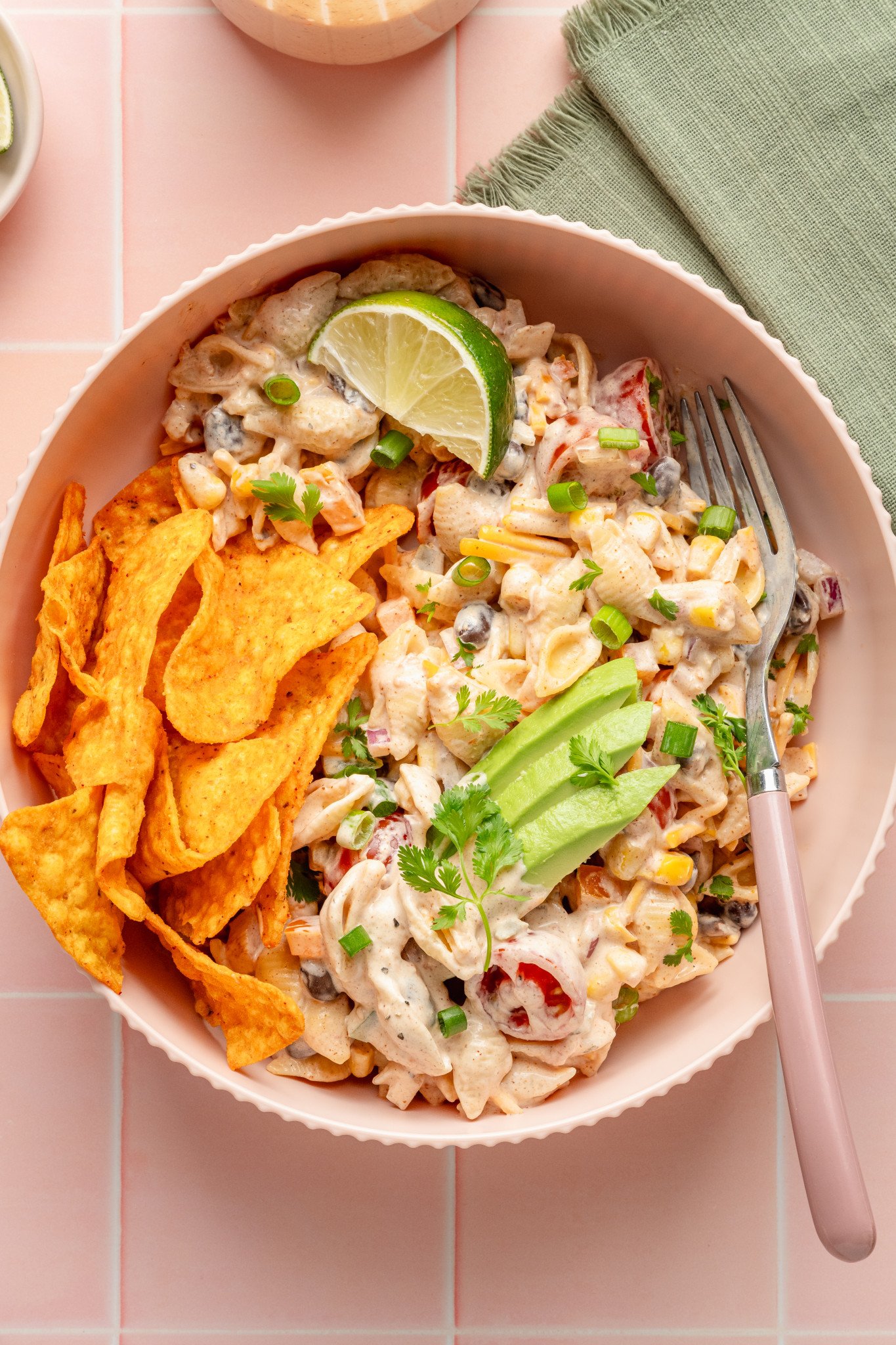 Close-up of creamy taco pasta salad served in a pink bowl, topped with avocado, cilantro, green onions, and a lime wedge. A portion of Doritos chips sits on the side, and a fork rests in the bowl.