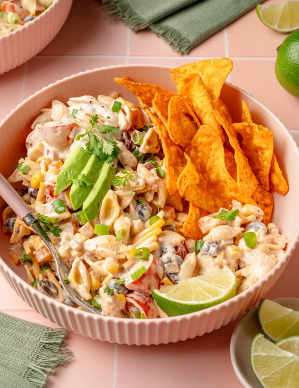Front-facing view of a bowl of creamy taco pasta salad topped with avocado slices, fresh cilantro, and green onions. A pile of Doritos chips is nestled beside the salad, and a lime wedge is placed in front for garnish.