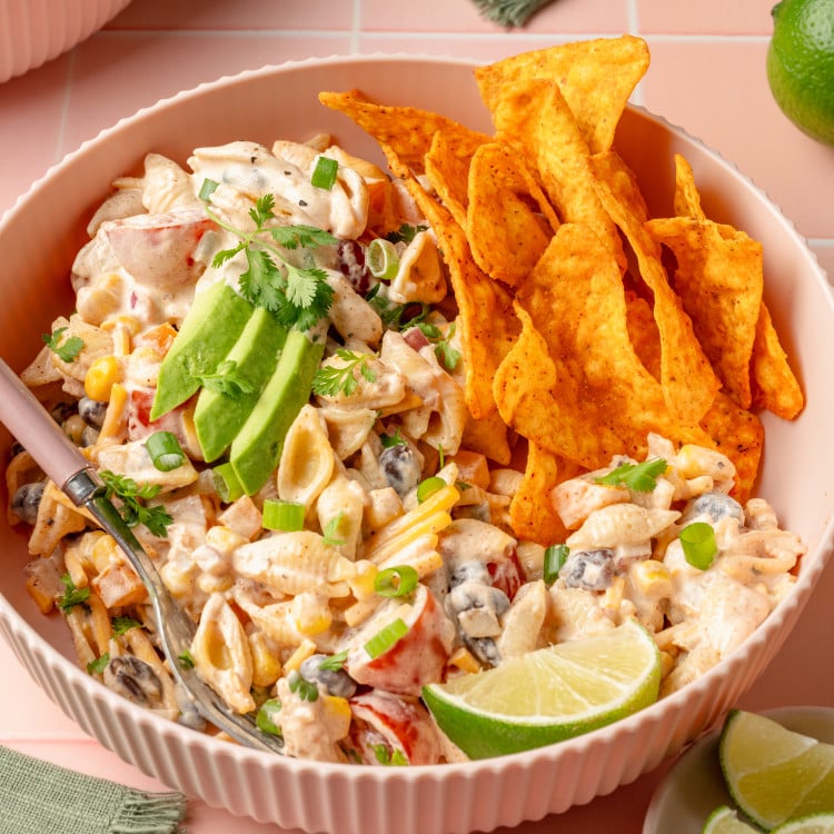 Front-facing view of a bowl of creamy taco pasta salad topped with avocado slices, fresh cilantro, and green onions. A pile of Doritos chips is nestled beside the salad, and a lime wedge is placed in front for garnish.