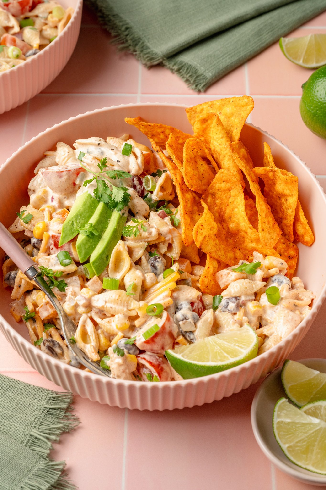 Front-facing view of a bowl of creamy taco pasta salad topped with avocado slices, fresh cilantro, and green onions. A pile of Doritos chips is nestled beside the salad, and a lime wedge is placed in front for garnish.
