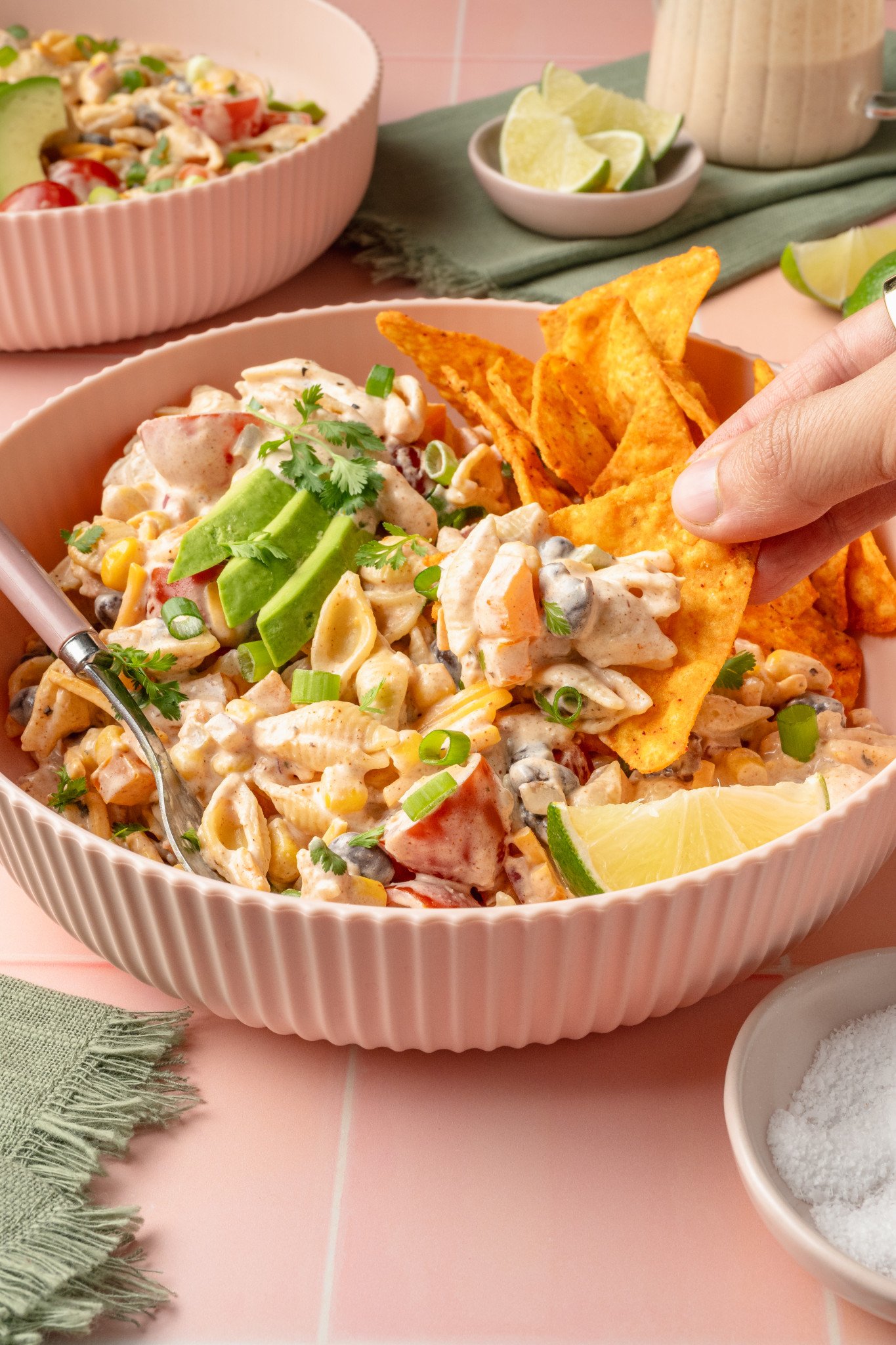 Side view of a hand holding a nacho chip topped with creamy taco pasta salad from a bowl garnished with avocado, cilantro, and lime wedges. The background includes another bowl of salad and a small bowl of salt.