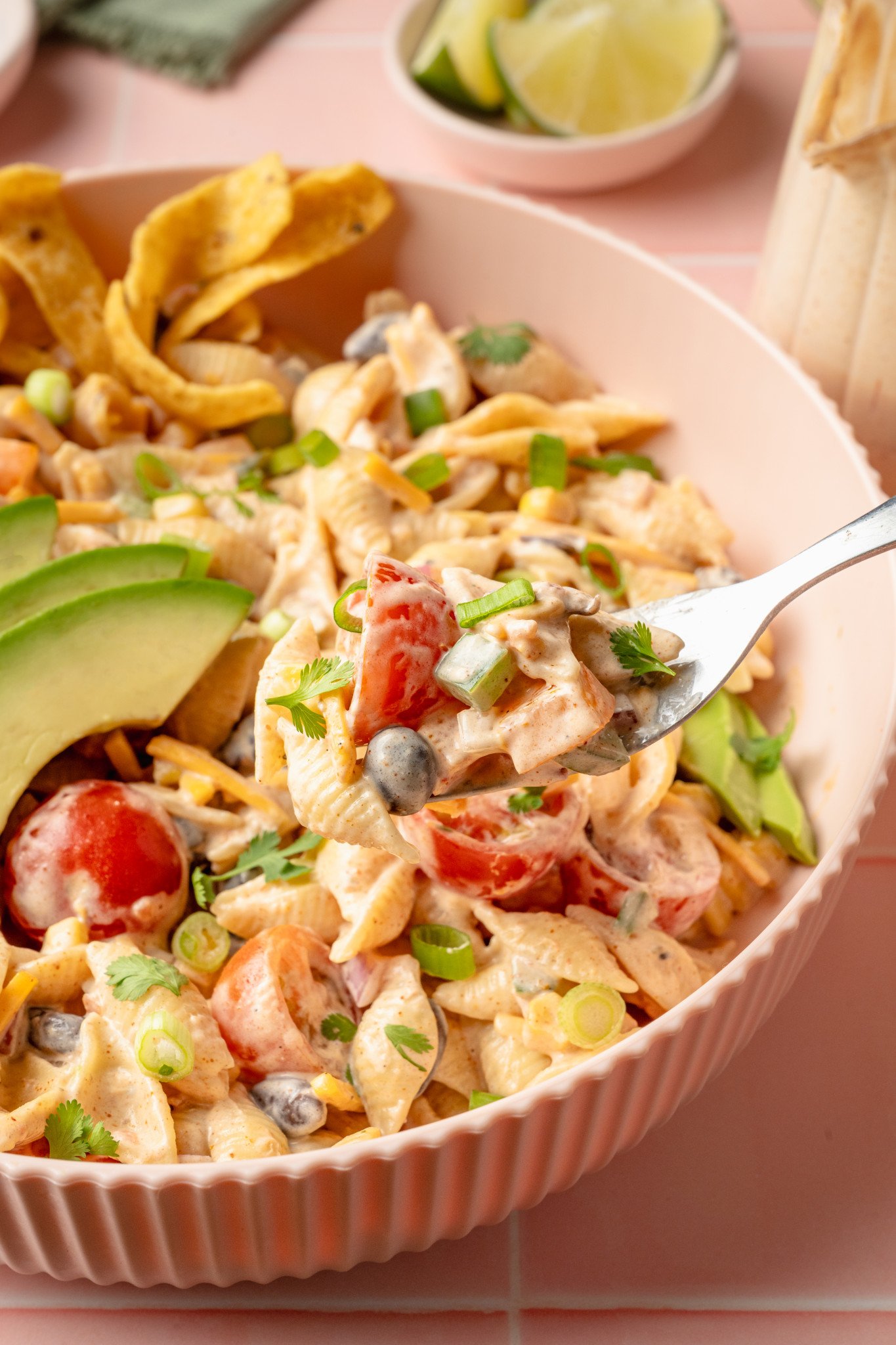 Close-up of a forkful of creamy taco pasta salad being lifted from a bowl. The fork holds shell pasta, cherry tomatoes, black beans, green onions, and bits of cilantro, with avocado and chips visible in the background.