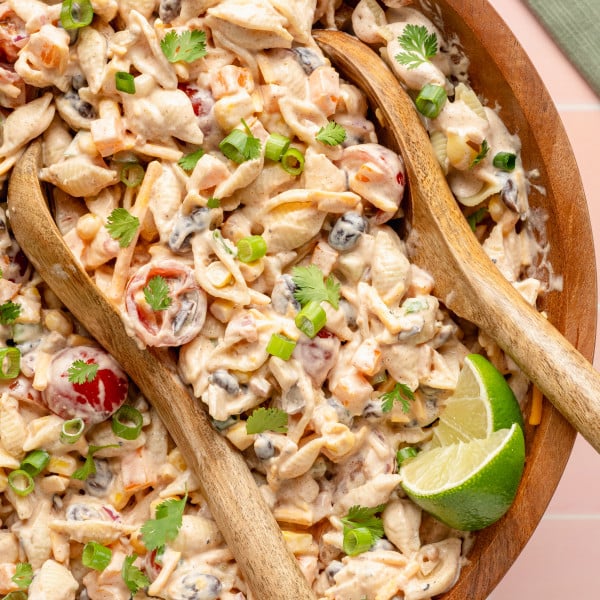 Close-up overhead shot of creamy taco pasta salad in a large wooden bowl with wooden serving spoons. The salad includes shell pasta, cherry tomatoes, corn, black beans, cheese, and is topped with green onions, cilantro, and a lime wedge.