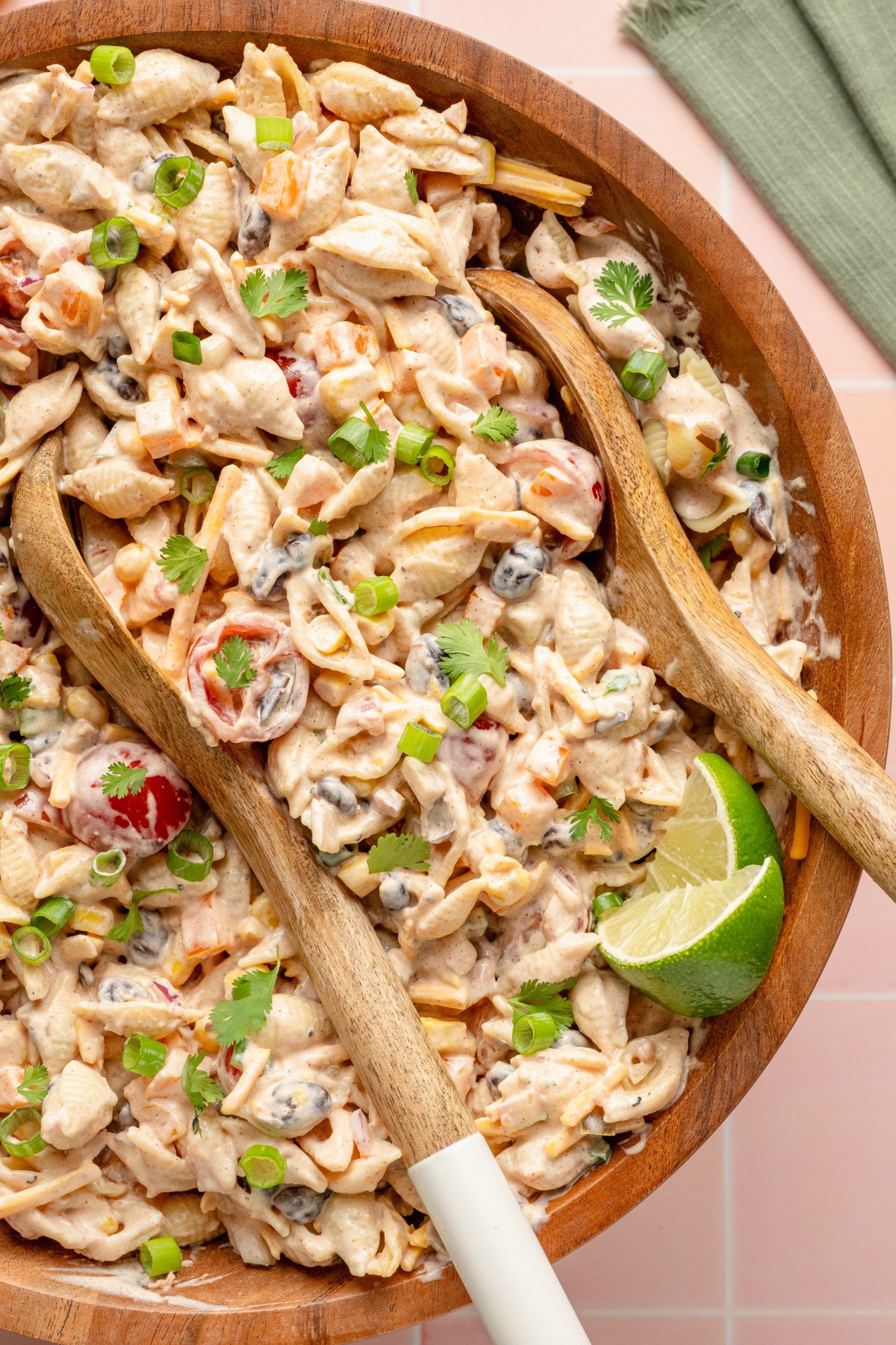 Close-up overhead shot of creamy taco pasta salad in a large wooden bowl with wooden serving spoons. The salad includes shell pasta, cherry tomatoes, corn, black beans, cheese, and is topped with green onions, cilantro, and a lime wedge.