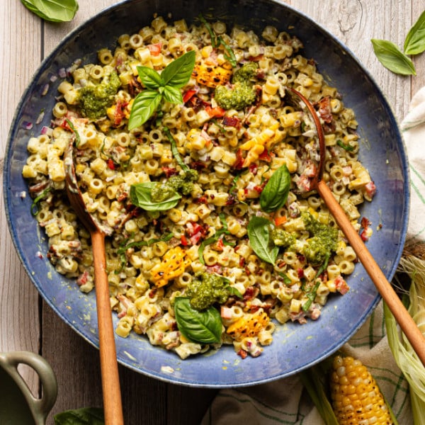 Pesto pasta salad tossed together in large blue serving bowl with wood spoons and garnished with fresh basil.