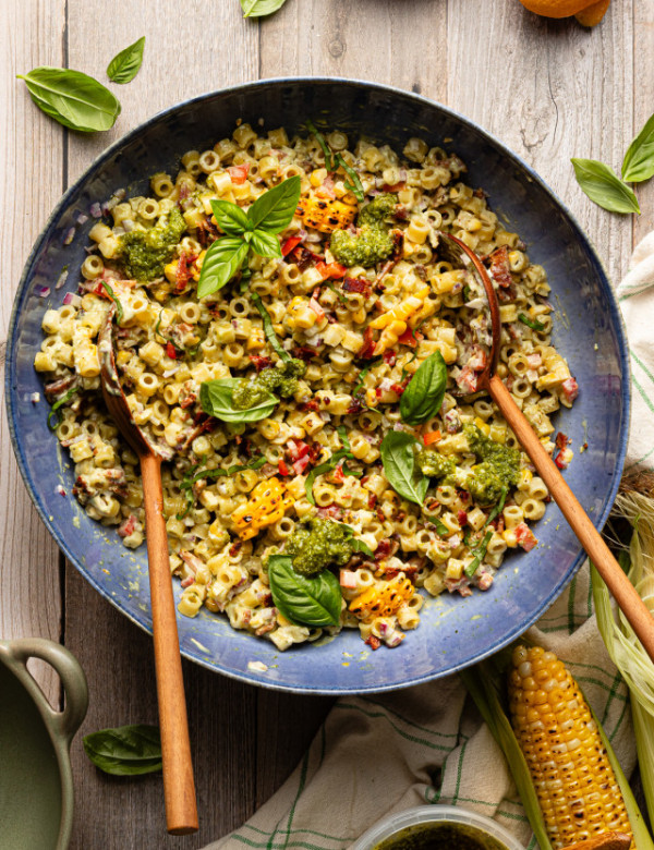 Pesto pasta salad tossed together in large blue serving bowl with wood spoons and garnished with fresh basil.