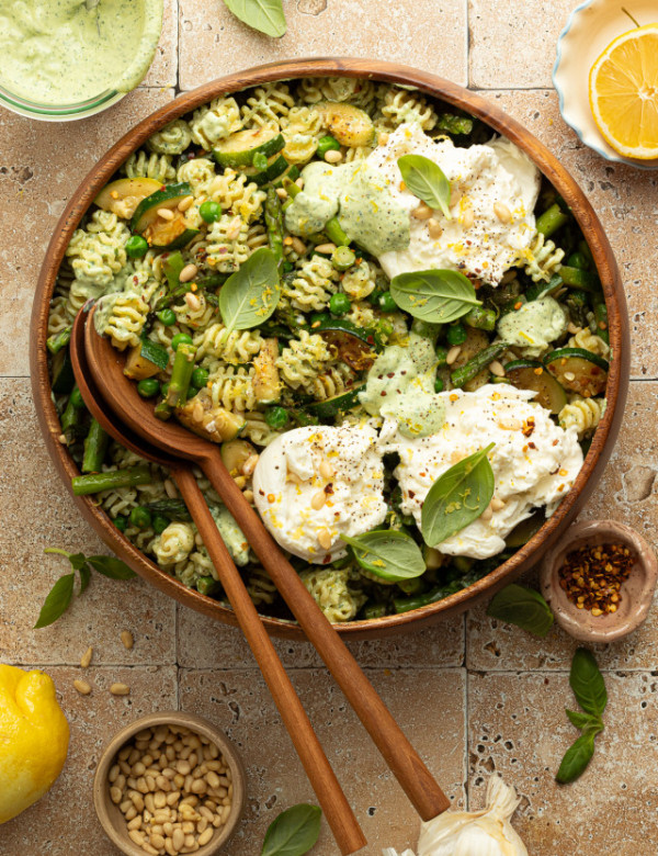 Overhead view of a finished green goddess pasta salad topped with dollops of burrata cheese, pine nuts, and fresh basil. The salad is served in a large wooden bowl surrounded by ingredients like lemon, red pepper flakes, and green herb dressing.