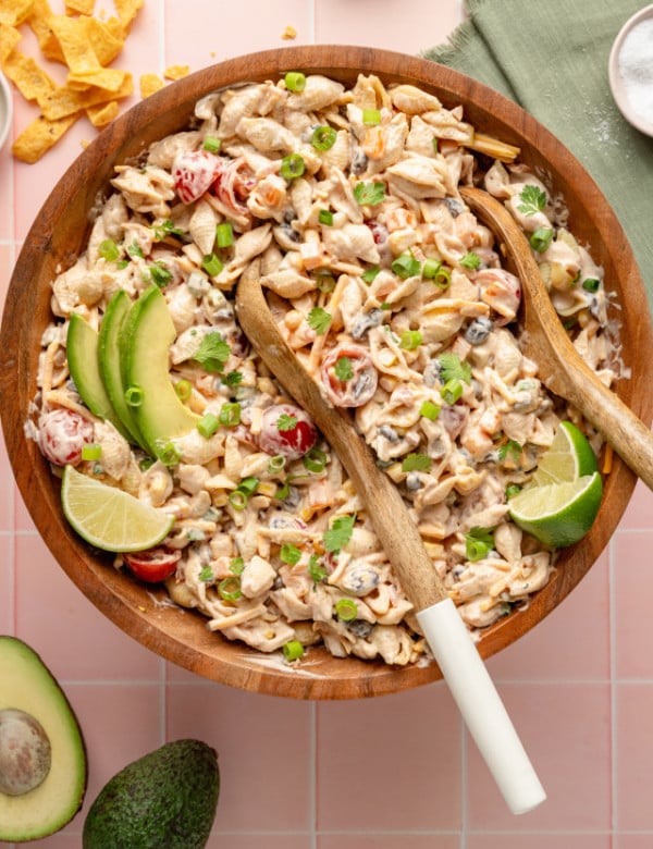 Overhead view of a large wooden bowl filled with creamy taco pasta salad, garnished with avocado slices, lime wedges, chopped green onions, and cilantro. Wooden serving utensils rest in the bowl, and taco chips are scattered around the pink tiled background.