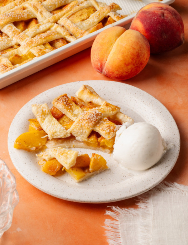 Partially eaten slice of peach slab pie with chunks of peach spilling out beside a melting scoop of ice cream. The pie dish and whole peaches are in the background.